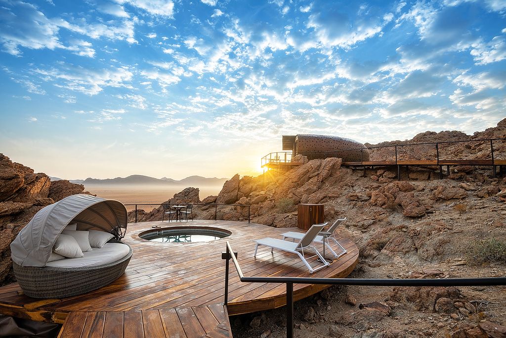 Private wooden deck with plunge pool and lounge chair overlooking vast Namibian desert landscape at golden sunrise.