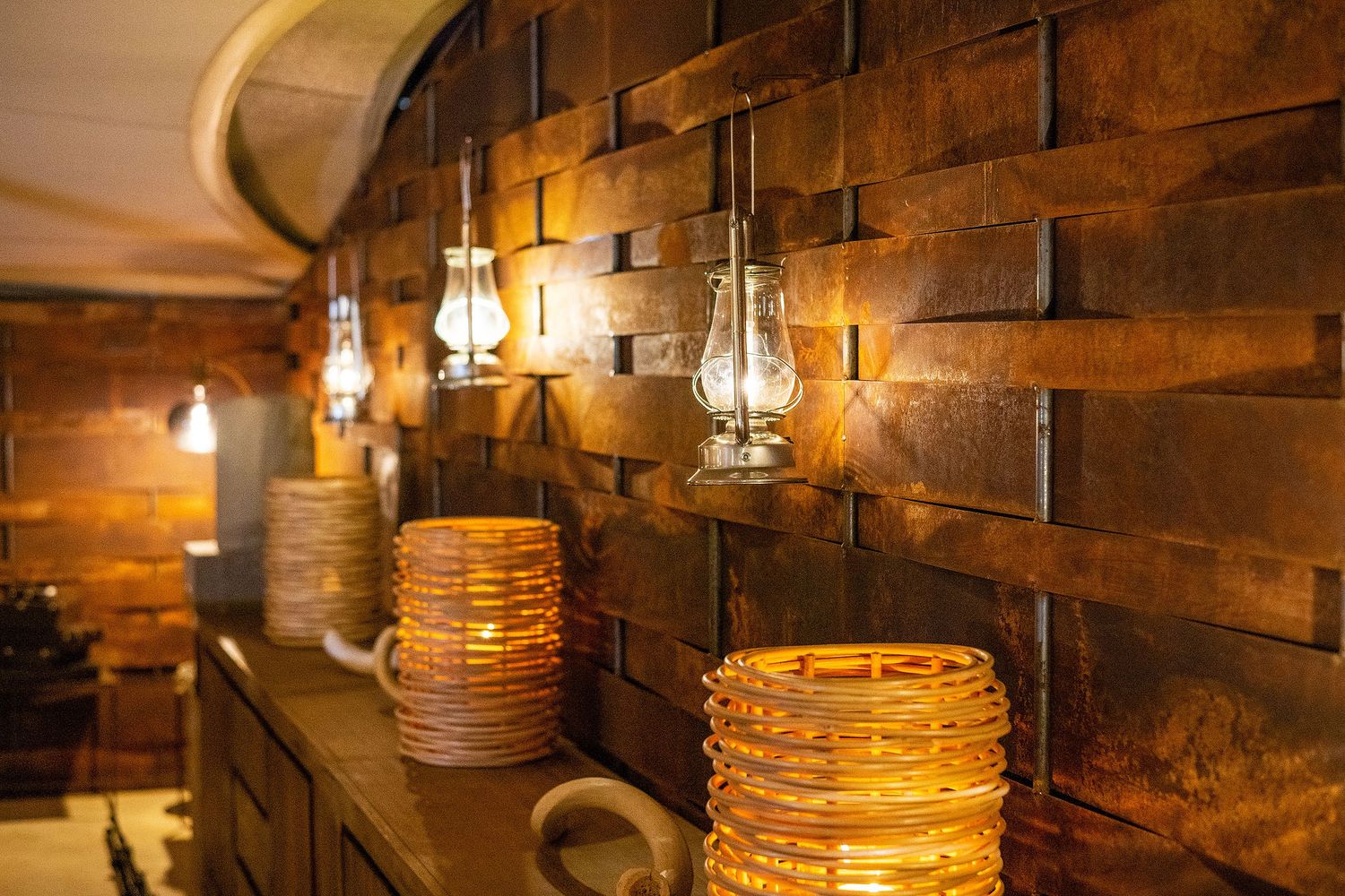 Rustic wooden wall with vintage oil lamp sconces illuminating woven rattan lanterns on a wooden shelf.