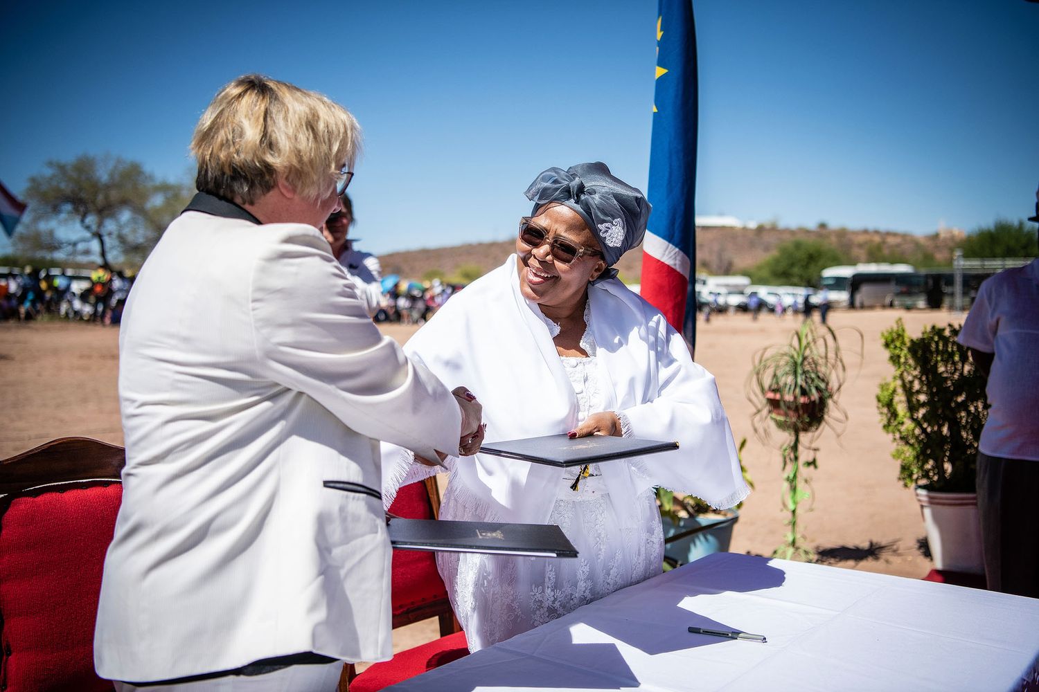 Two women shaking hands at an outdoor ceremony with EU and Namibia flags in the background.
