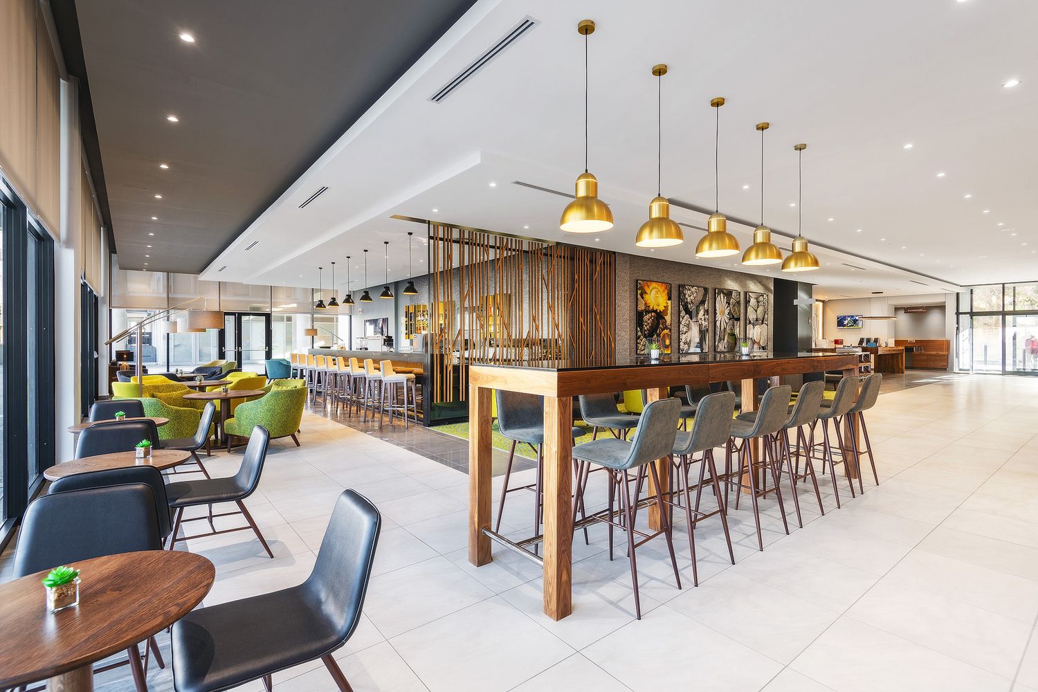 Modern hotel bar and lounge with gold pendant lights, green chairs, and a sleek wooden bar counter.