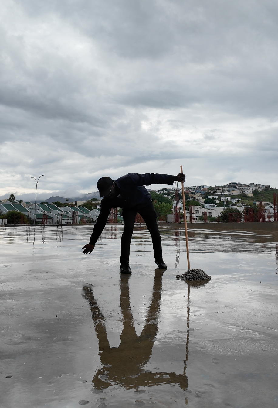 Silhouetted worker mopping a wet reflective surface outdoors under cloudy skies with hillside buildings in background.
