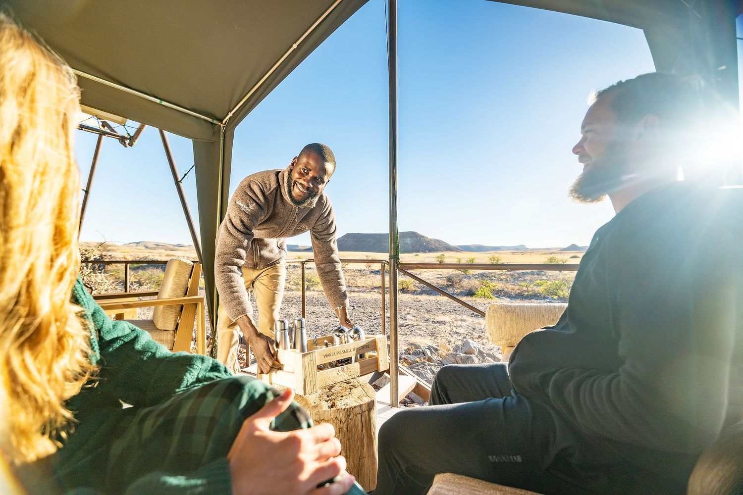 Safari guide serving drinks to tourists in an open-air game drive vehicle in a sunny African savanna landscape.