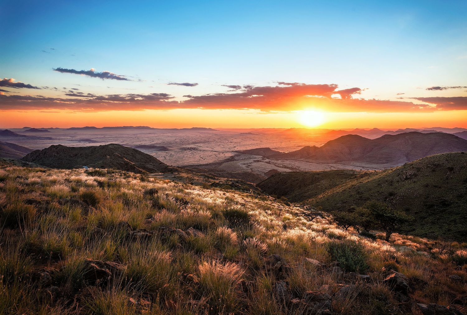 Golden sunset over rolling mountains with dry grasslands in the foreground and a vast valley below.