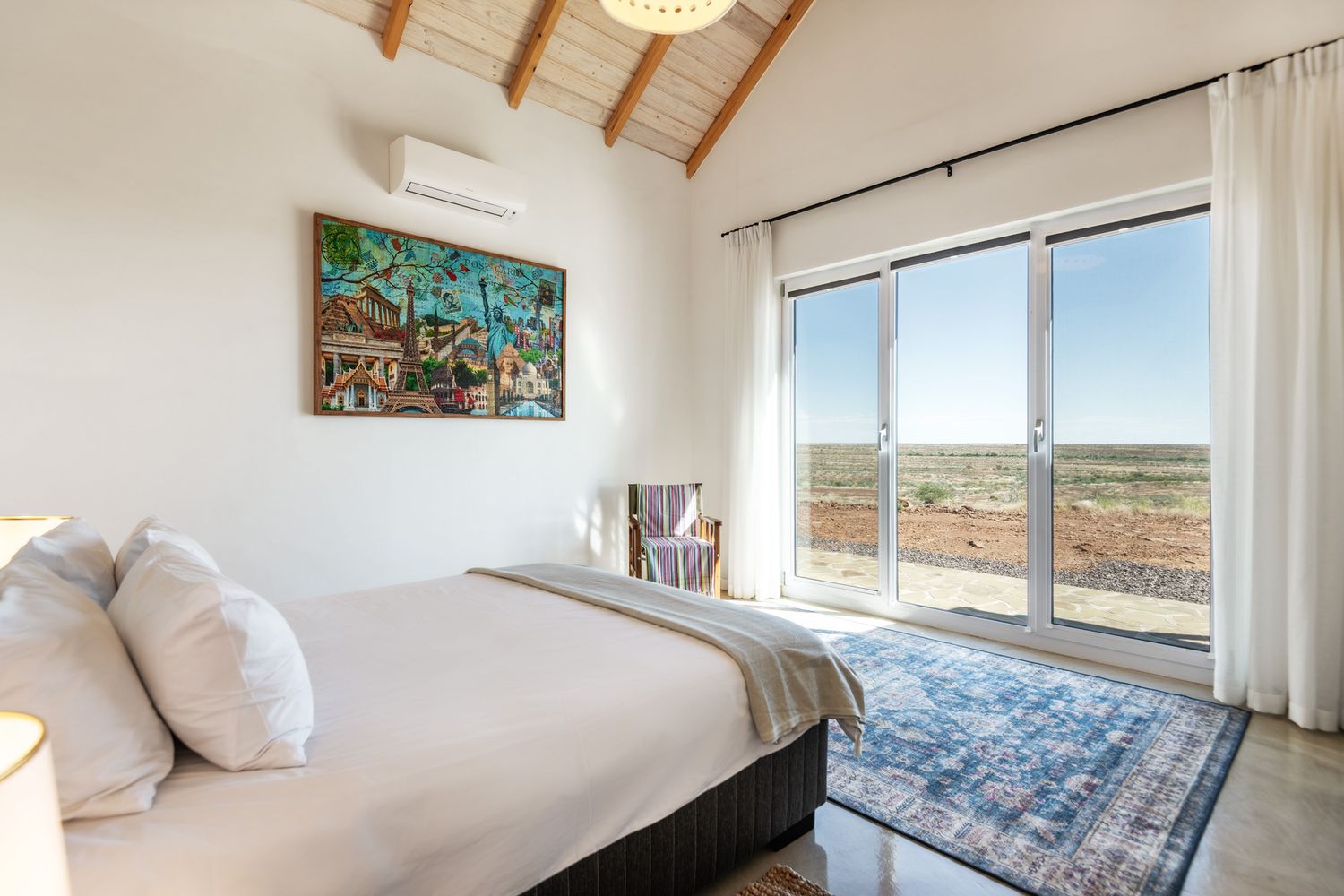 Bright bedroom with white bedding, wood-beam ceiling, wall AC unit, colorful artwork, and large glass doors overlooking arid landscape.