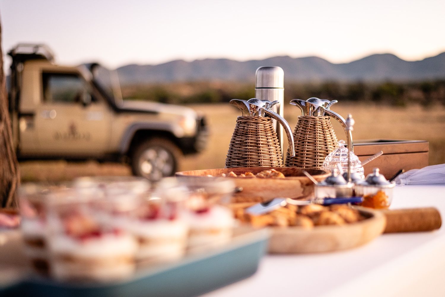 Safari sundowner spread with wicker decanters, snacks on wooden boards, and a 4x4 vehicle in the African savanna background.