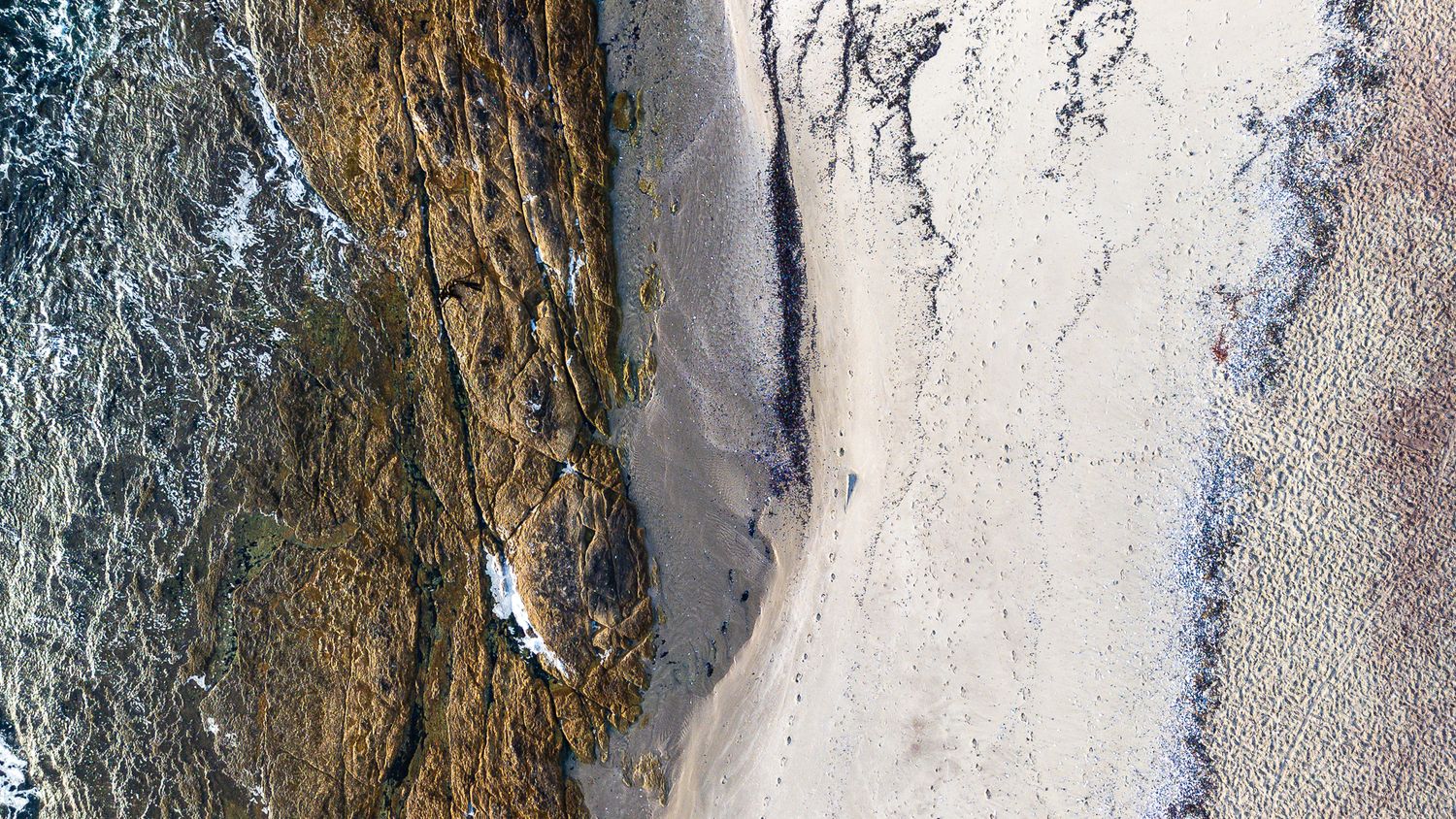 Aerial view of rocky coastline meeting white sandy beach with gentle ocean waves.