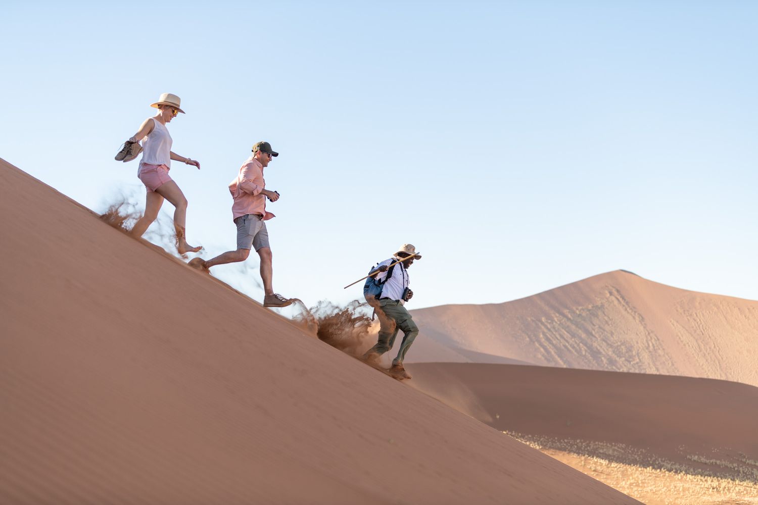 Three tourists hiking up a steep red sand dune in the Namib Desert under a clear blue sky.