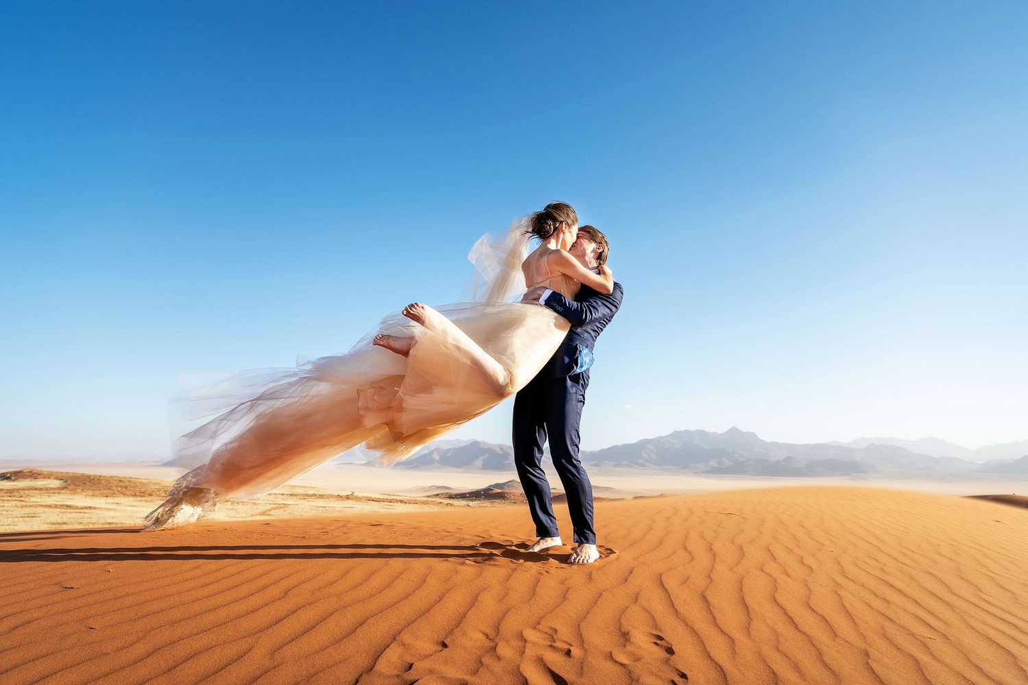 Groom lifting bride in white wedding dress on golden sand dunes under clear blue sky in desert landscape.
