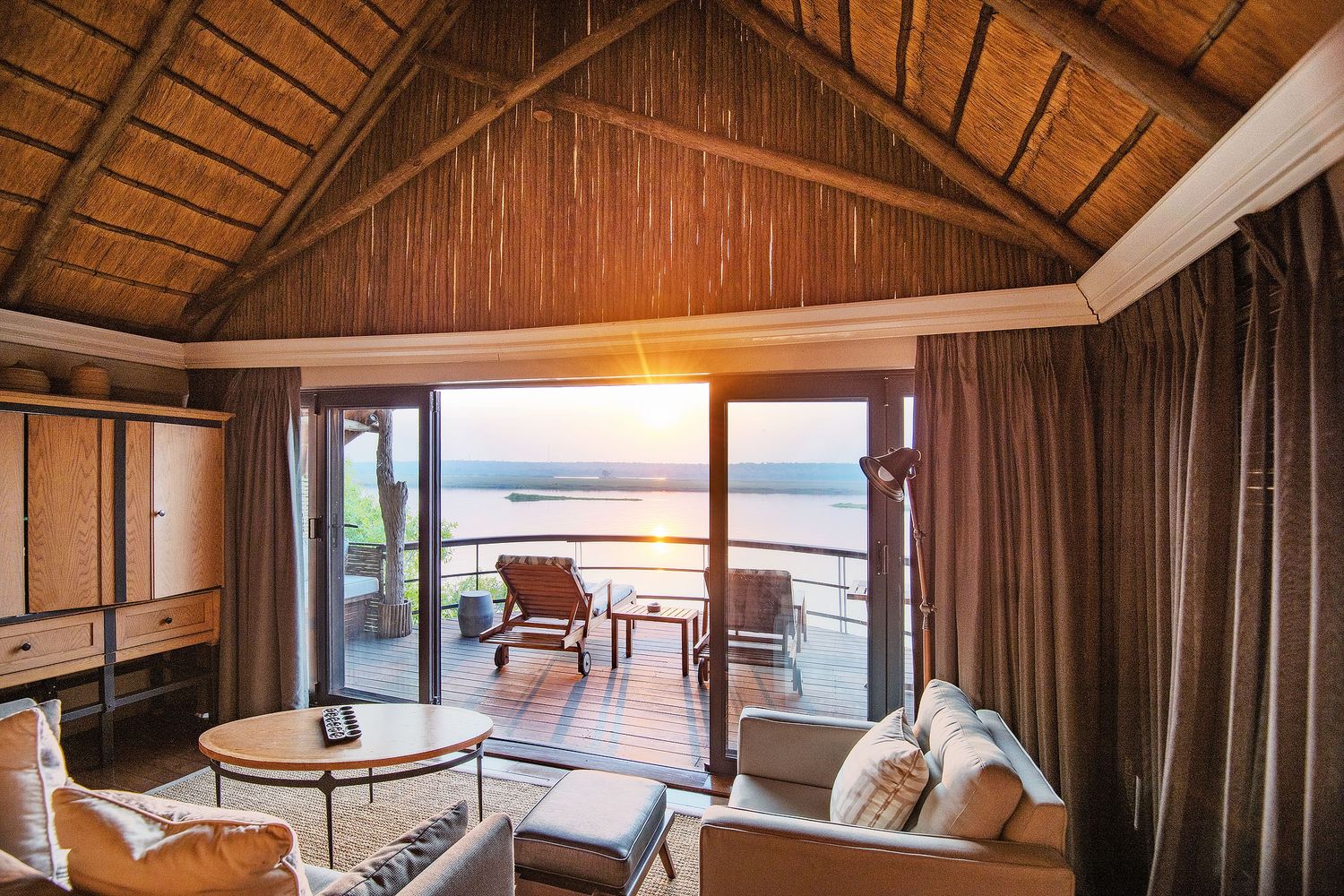 Luxury safari suite interior with thatched roof, elegant furnishings, and panoramic deck views of African sunset.