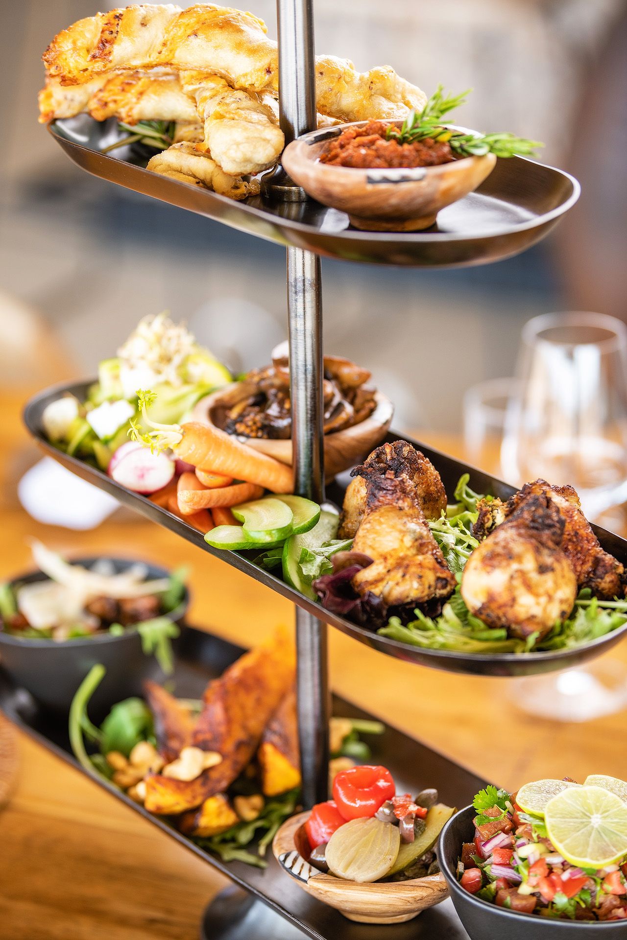 A three-tiered serving stand filled with assorted appetizers, grilled chicken, fresh salads, and dipping sauces at a restaurant.