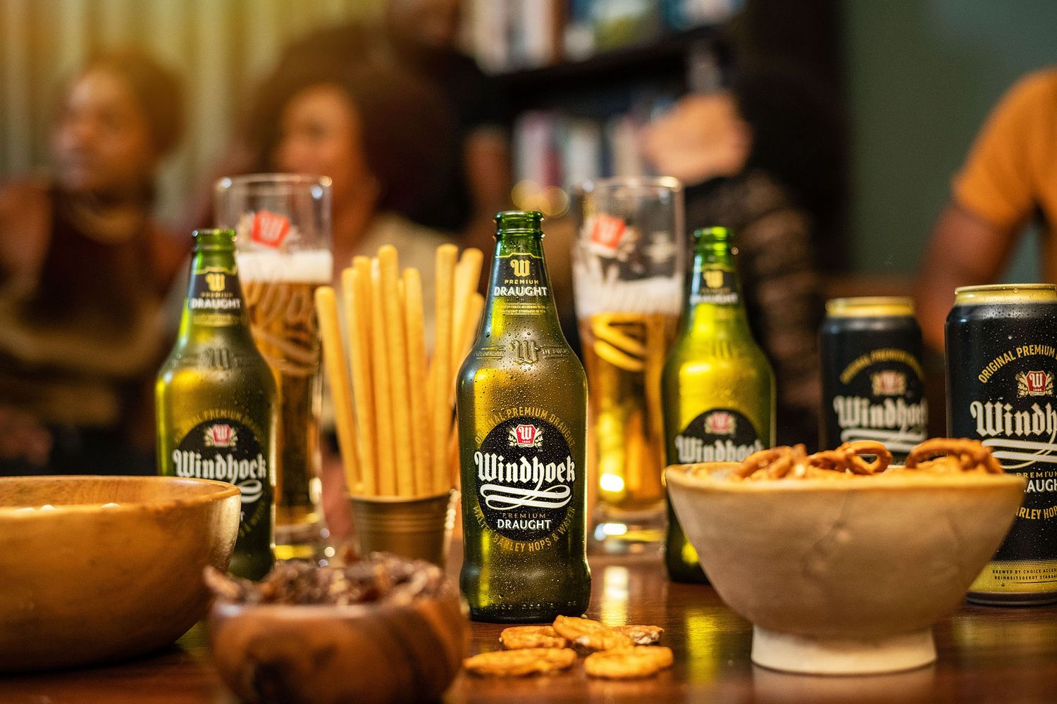 Windhoek lager beer bottles on a table with snacks and blurred people socializing in the background.