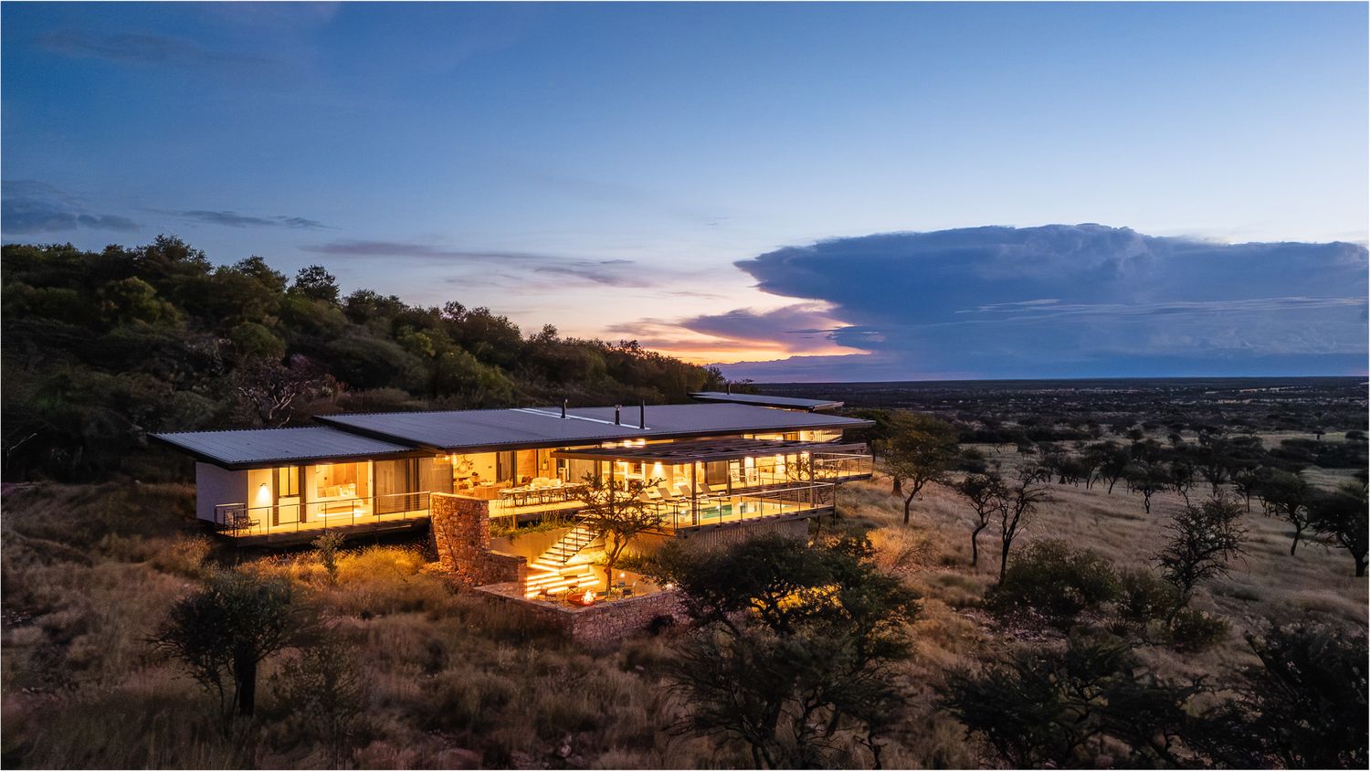 Contemporary glass and steel safari lodge glowing at dusk amid vast African savanna grasslands.
