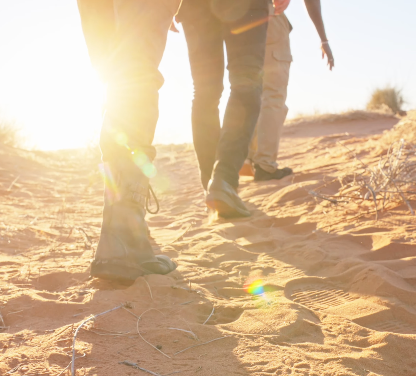Two people walking on red desert sand dunes at sunset with warm golden light flaring behind them.
