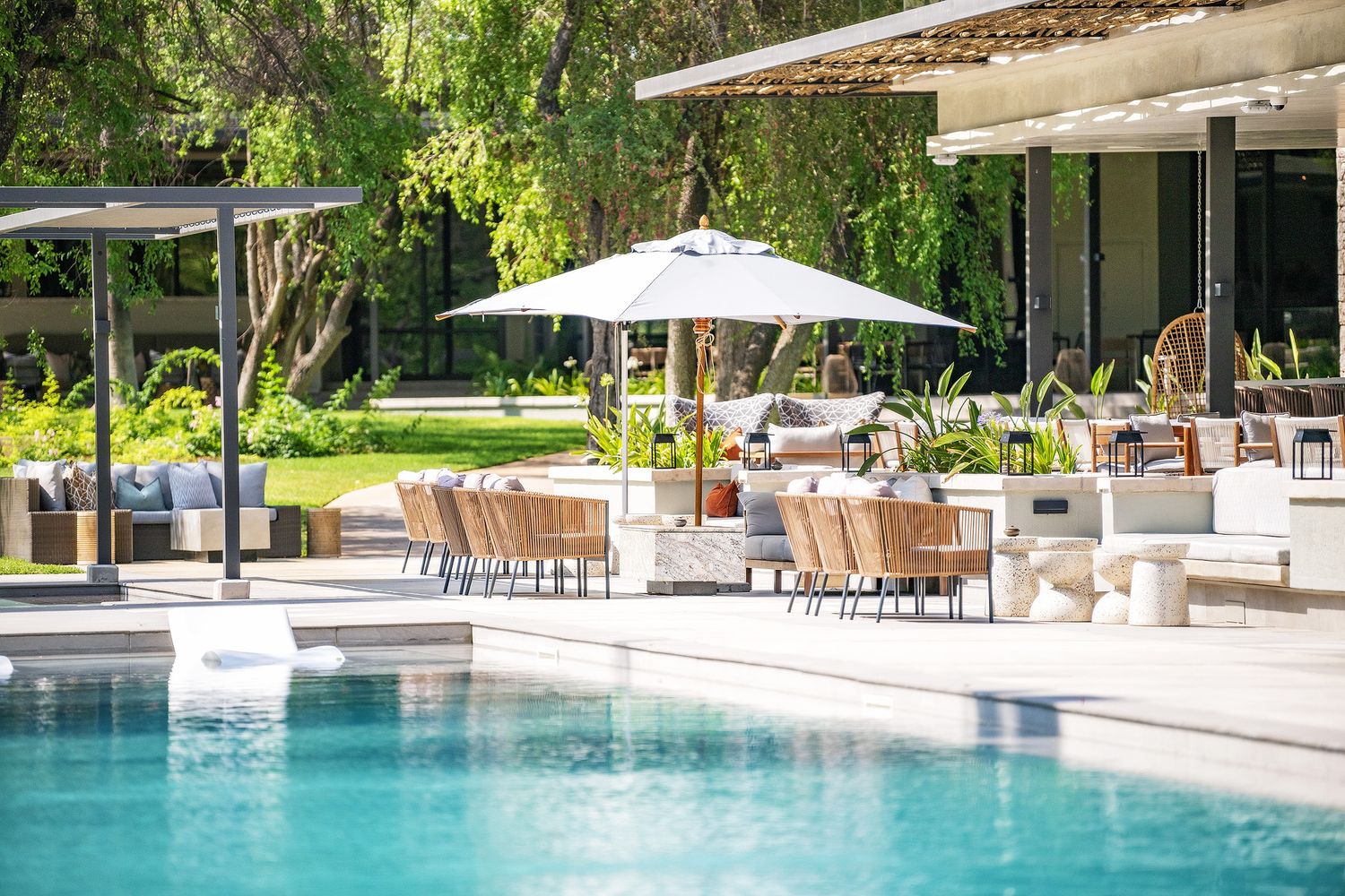 Luxury resort pool area with wicker chairs, white umbrellas, and lush greenery surrounding a crystal-clear blue swimming pool.