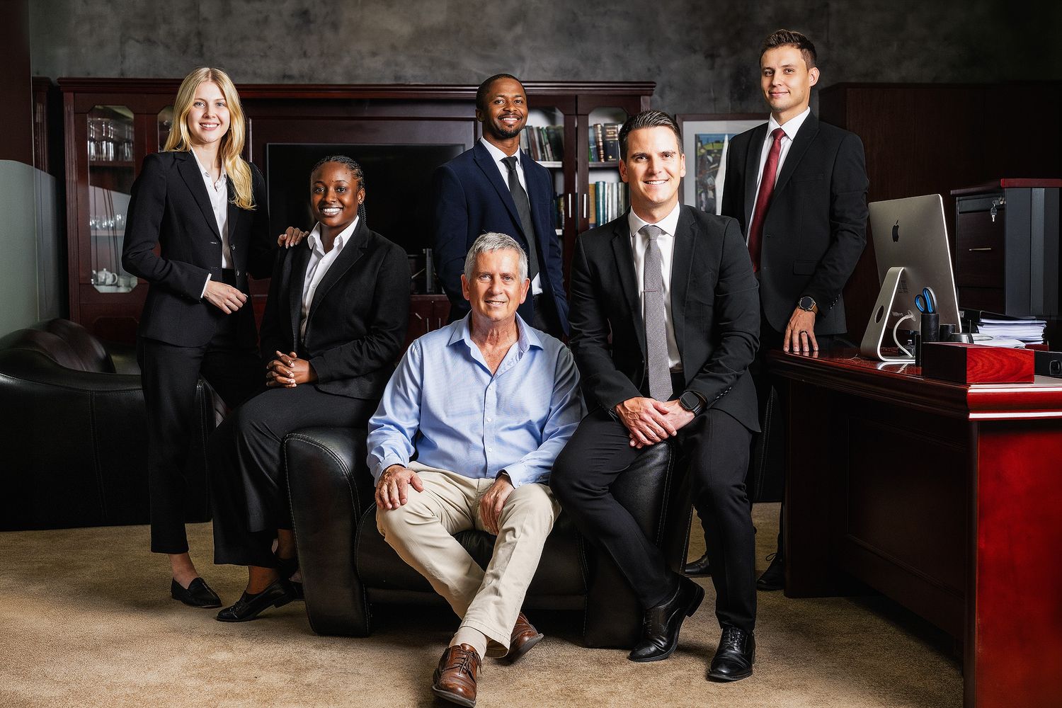 Six business professionals posing confidently in a formal office setting, with one seated and five standing behind him.