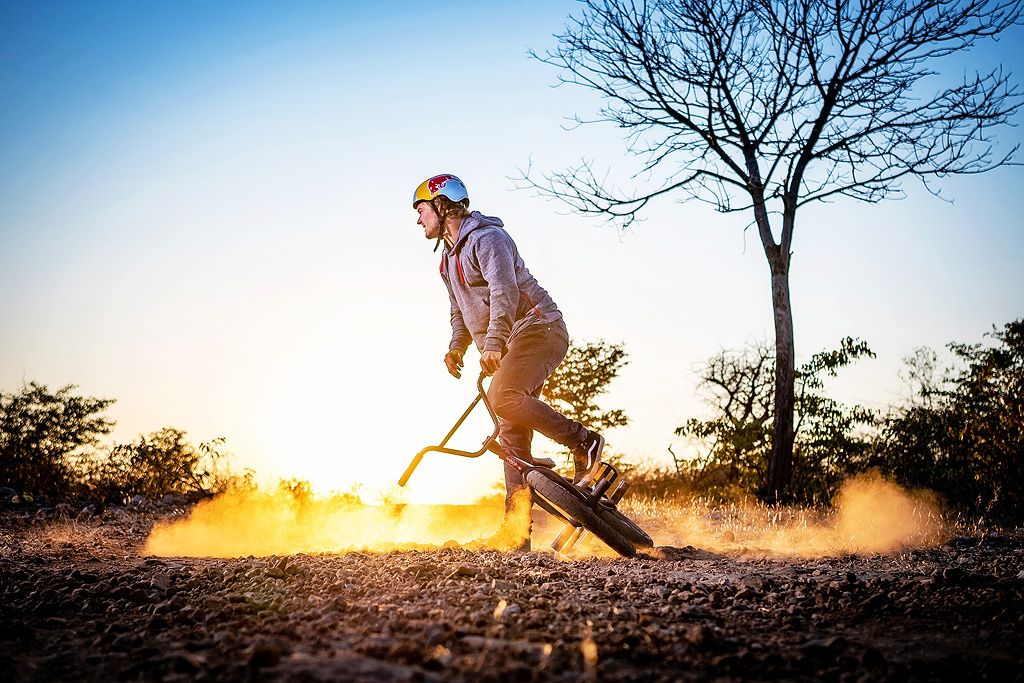 Person riding a scooter on dirt trail at sunset wearing a helmet, with golden light and dust clouds in background.