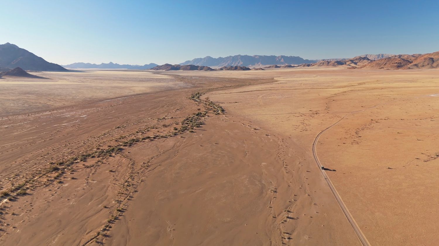 Aerial view of orange Namib Desert landscape with a lone vehicle on a desert road.
