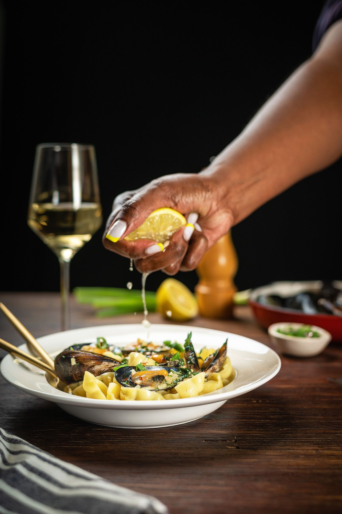 Hand squeezing fresh lemon over pasta with mussels and greens, served with white wine on rustic wooden table.