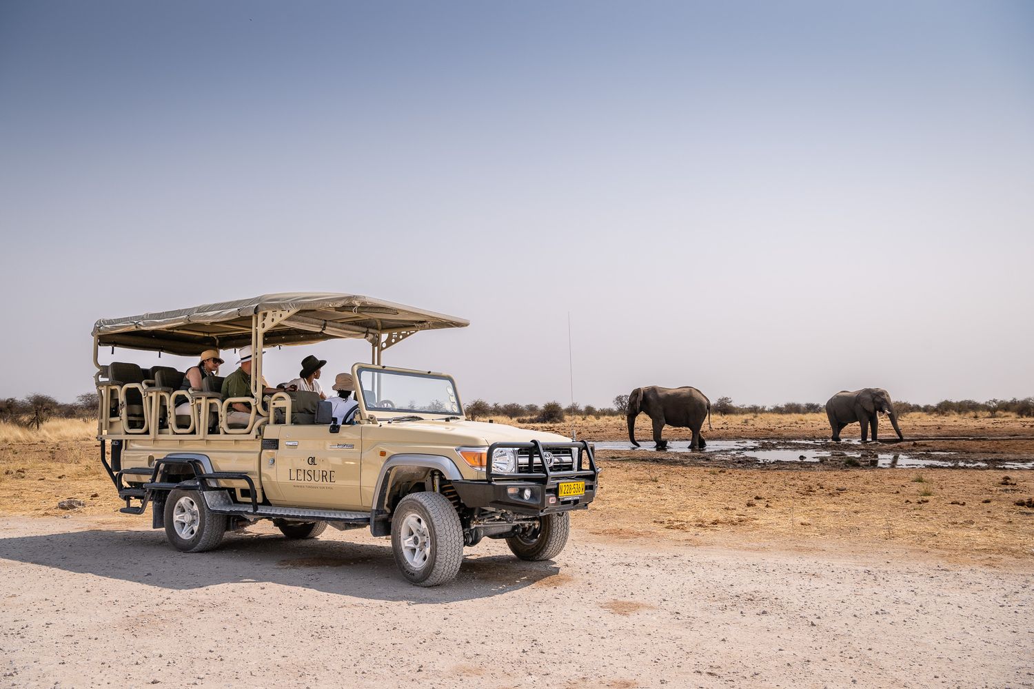 Safari guests observe wild elephants at waterhole from open Land Cruiser game drive vehicle in African bush.