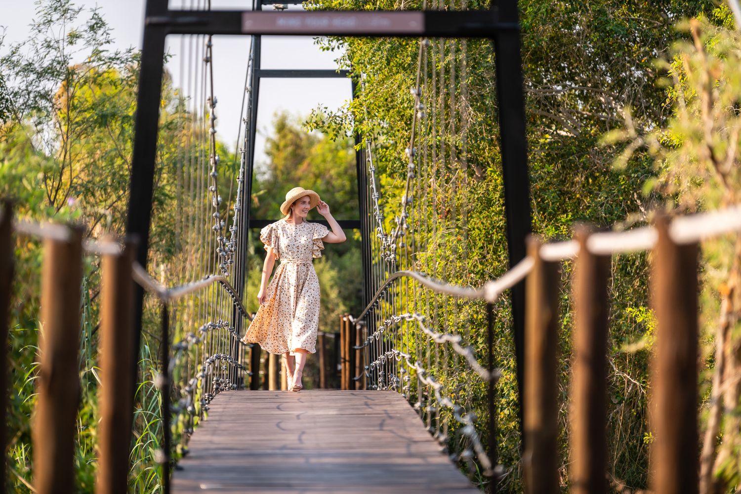Woman in floral dress walking on wooden suspension bridge surrounded by lush green trees.