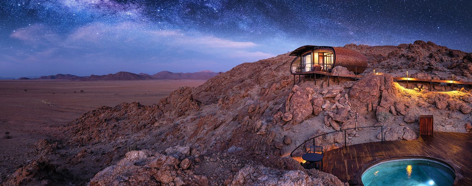 Starlit Milky Way panorama over a remote luxury desert lodge with illuminated plunge pool among rugged rocks.