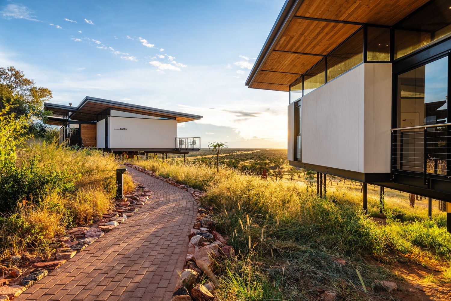 Modern lodge buildings connected by brick pathway surrounded by wild grasslands at golden sunset.