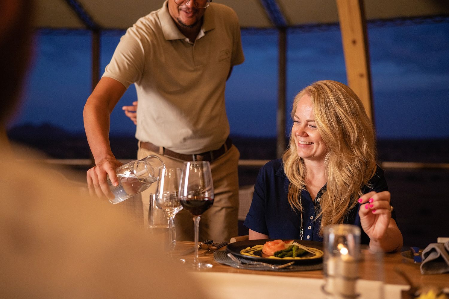 A smiling woman enjoys a fine dining experience with wine and gourmet food served by a waiter at dusk.