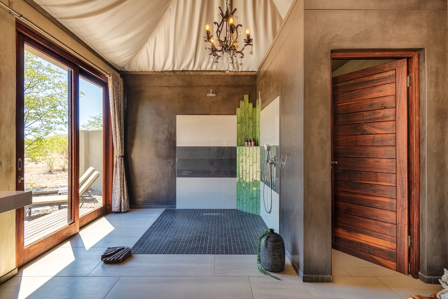 Luxurious open-air bathroom with concrete walls, wooden door, chandelier, walk-in shower, and large glass doors overlooking outdoor patio.