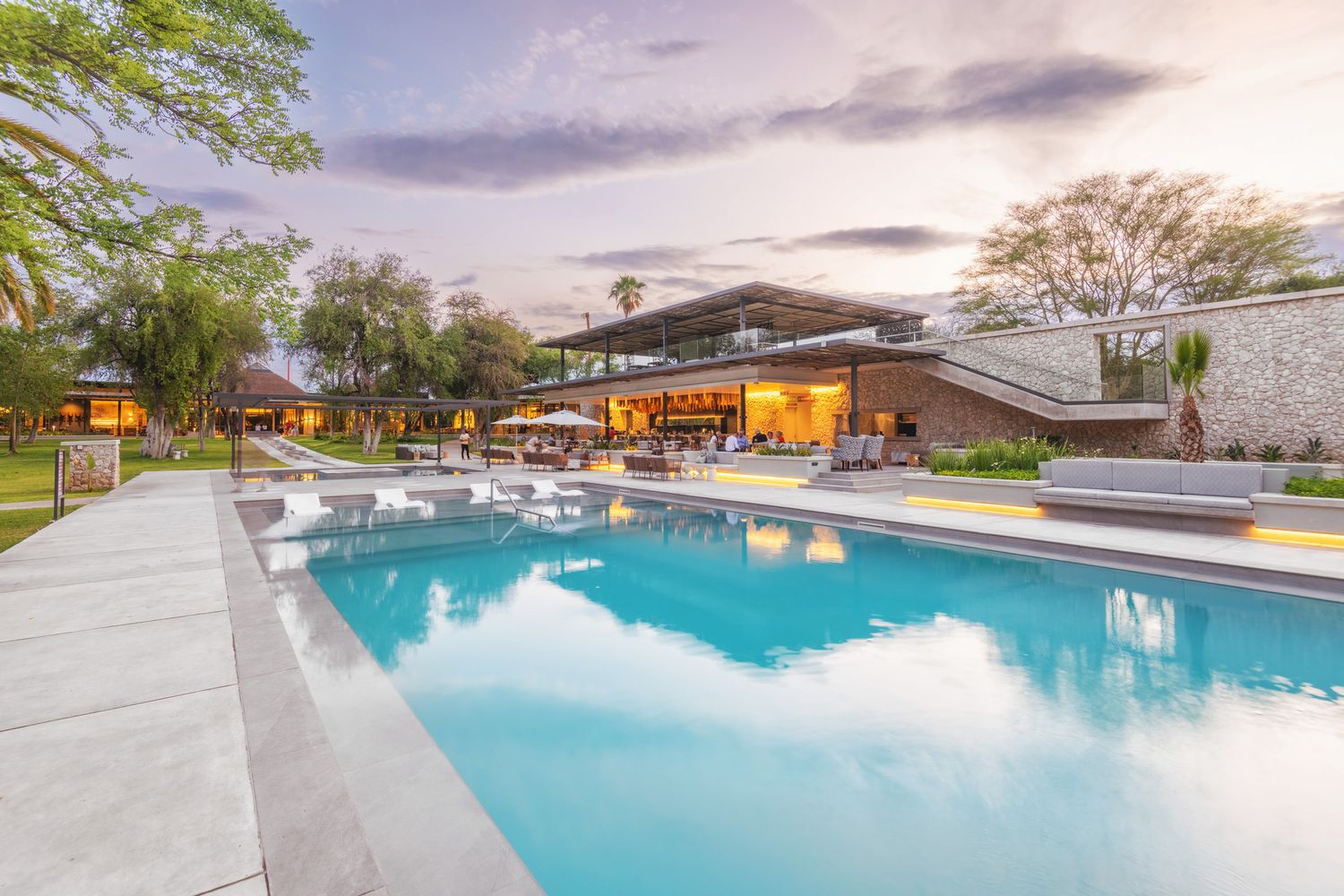 Luxury safari lodge with stunning infinity pool reflecting colorful sunset sky surrounded by lush tropical gardens.