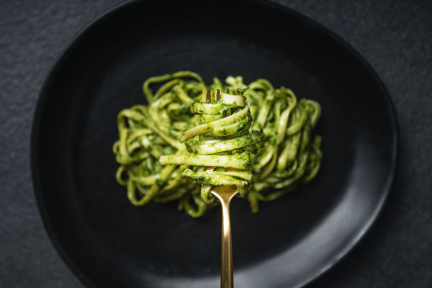 A fork twirled with green pesto fettuccine pasta served on a dark black plate, top-down view.