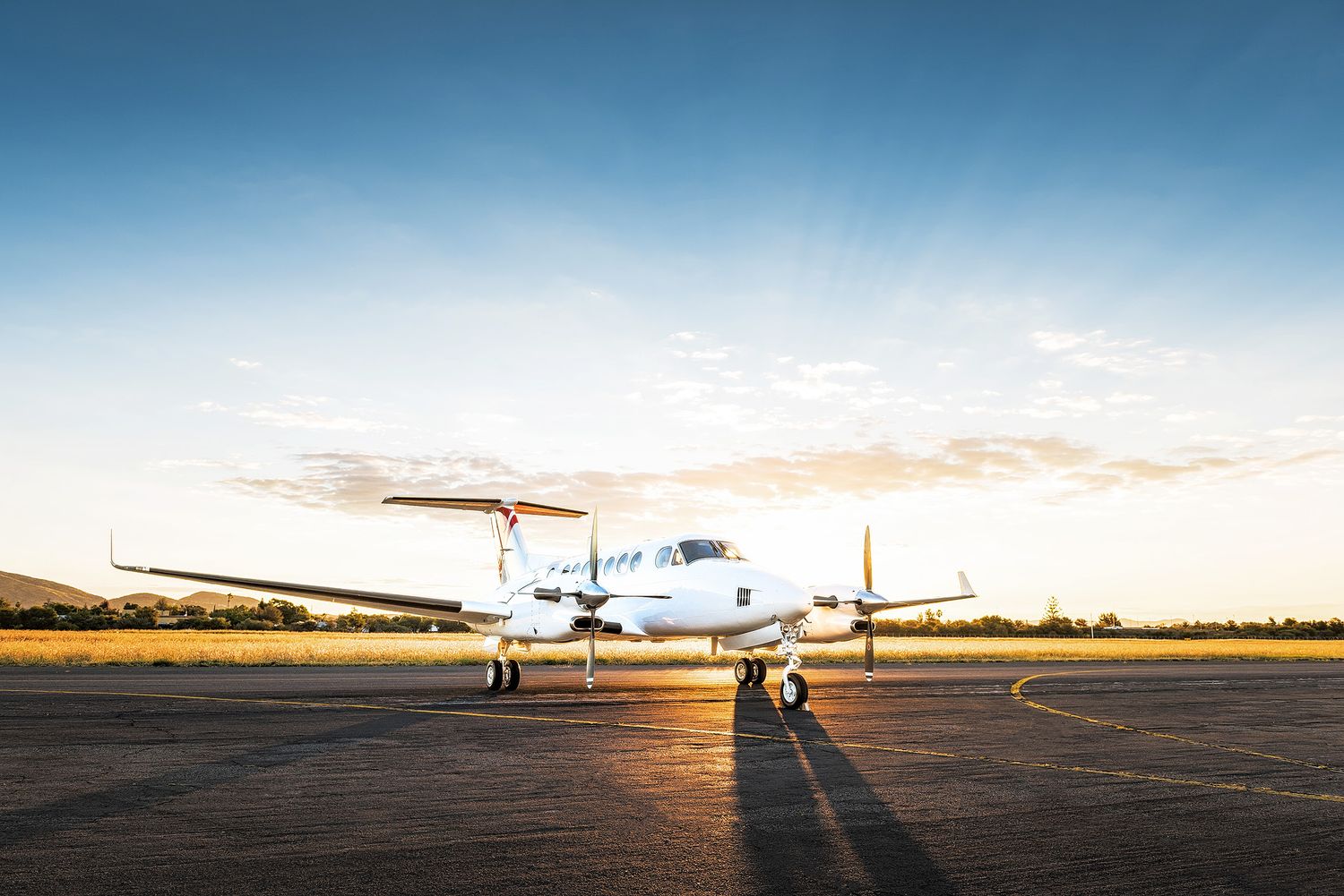 White turboprop aircraft parked on tarmac at golden hour sunset with clear blue sky and mountains in background.