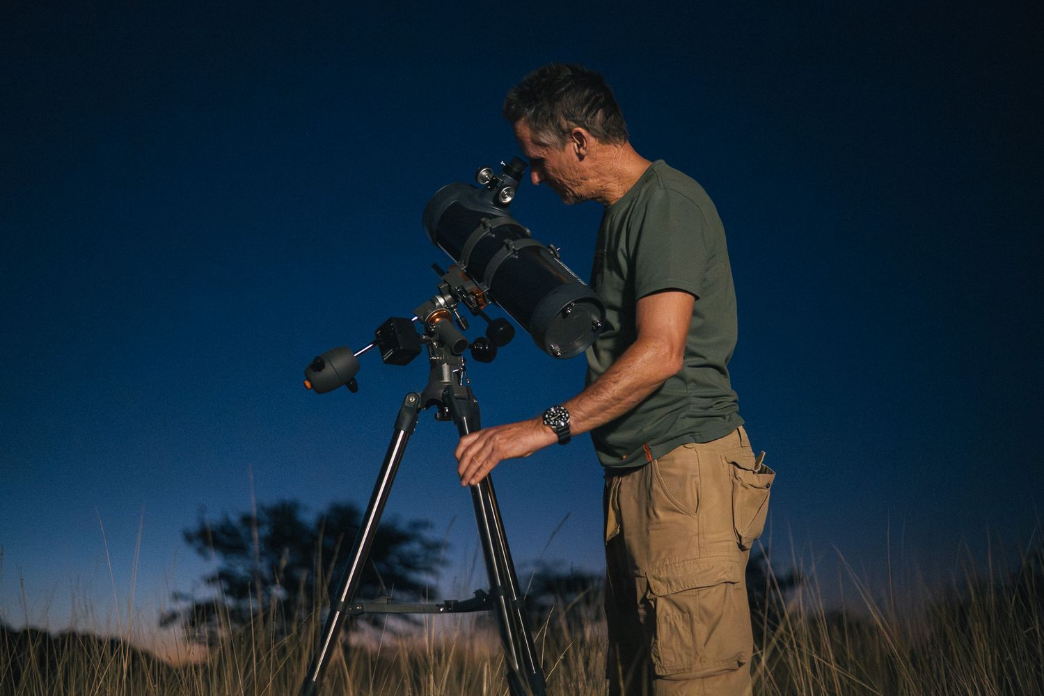 Man using a telescope outdoors at night under a deep blue sky, stargazing in a grassy field.