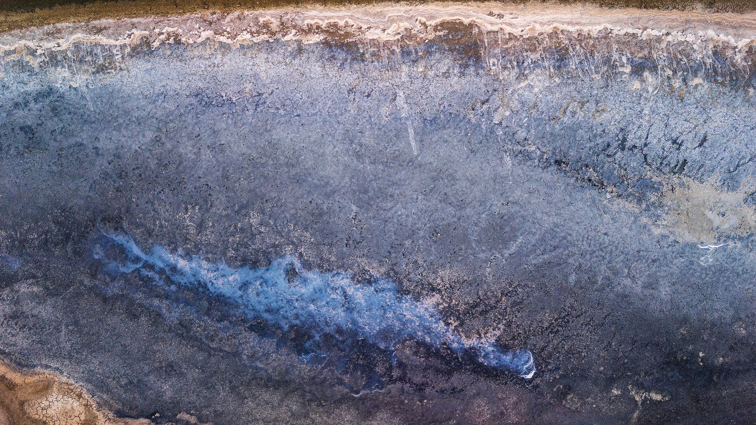 Aerial view of ocean waves crashing on a dark rocky shoreline with blue water and white seafoam patterns.