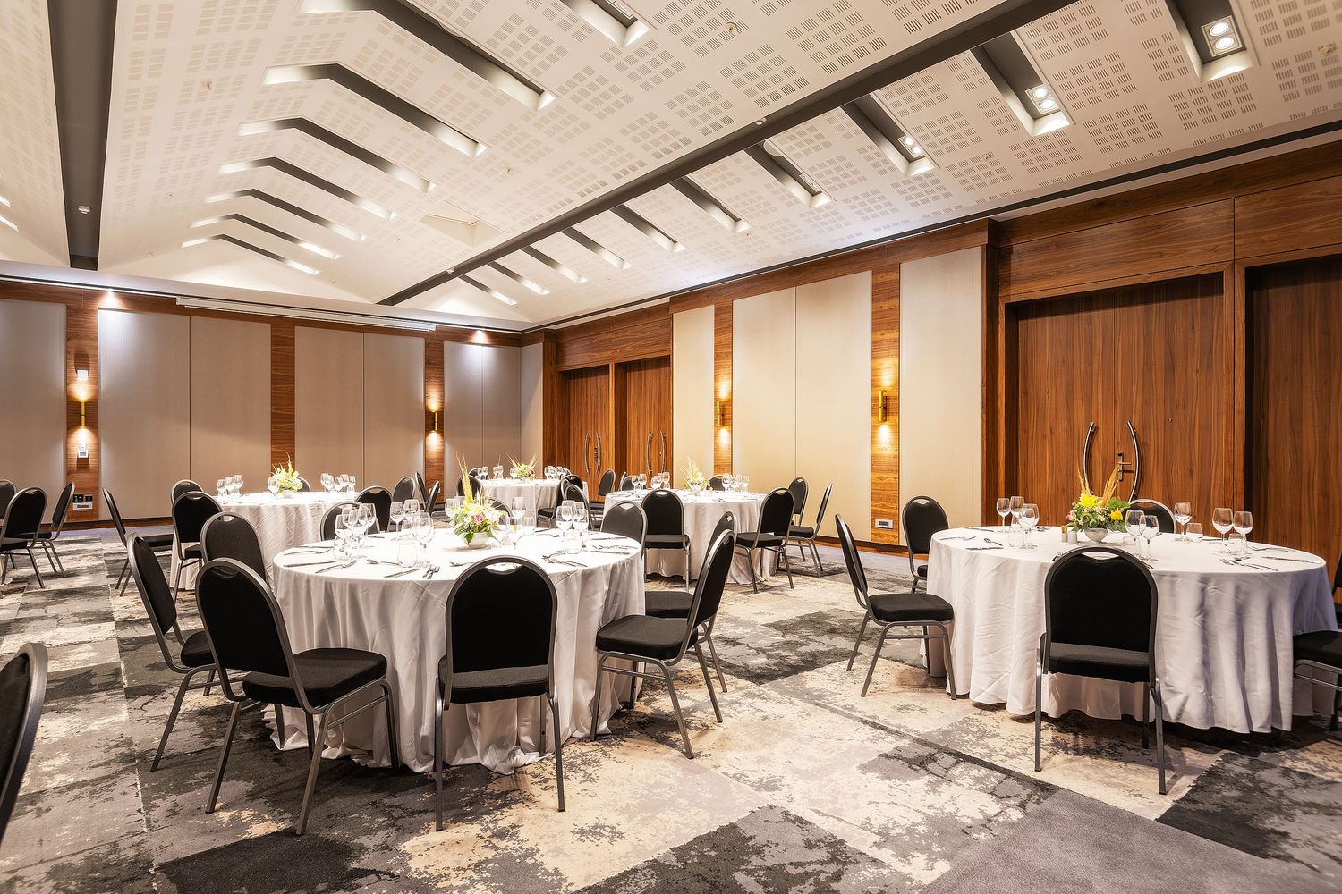 Elegant hotel banquet hall with round tables set for dining, black chairs, warm wood panels, and modern ceiling design.