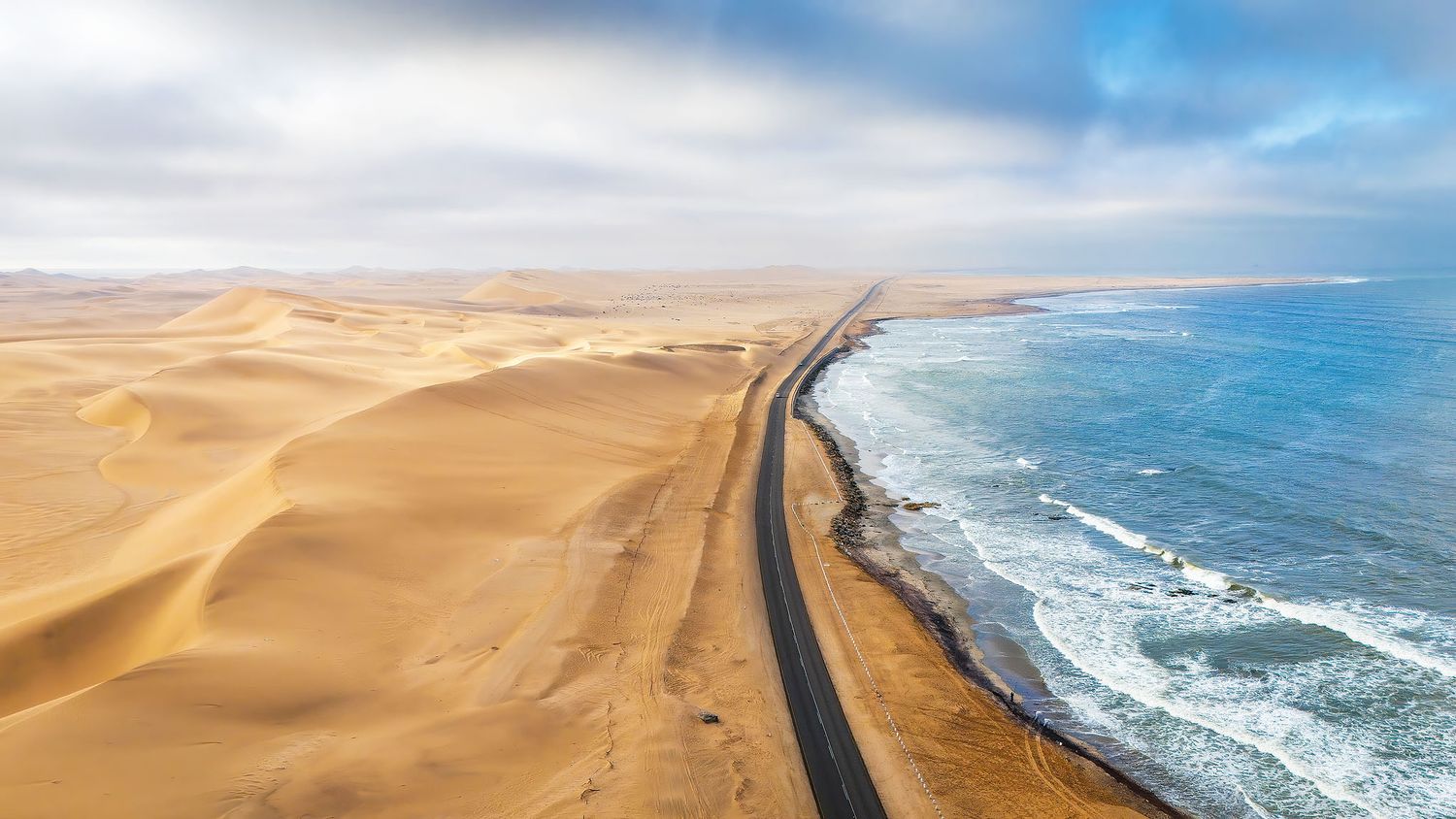 Aerial view of a desert road between golden sand dunes and the Atlantic Ocean coastline under a cloudy sky.