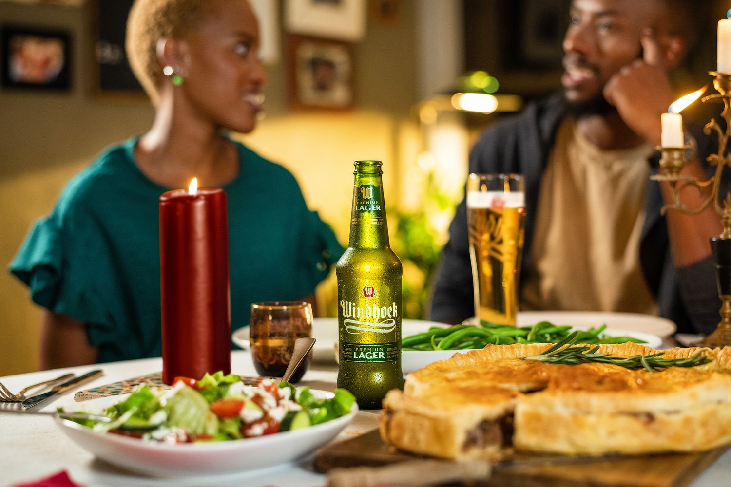 Two people dining at a candlelit table with Stellenbosch Lager beer, fresh salad, and pizza in a warm restaurant setting.