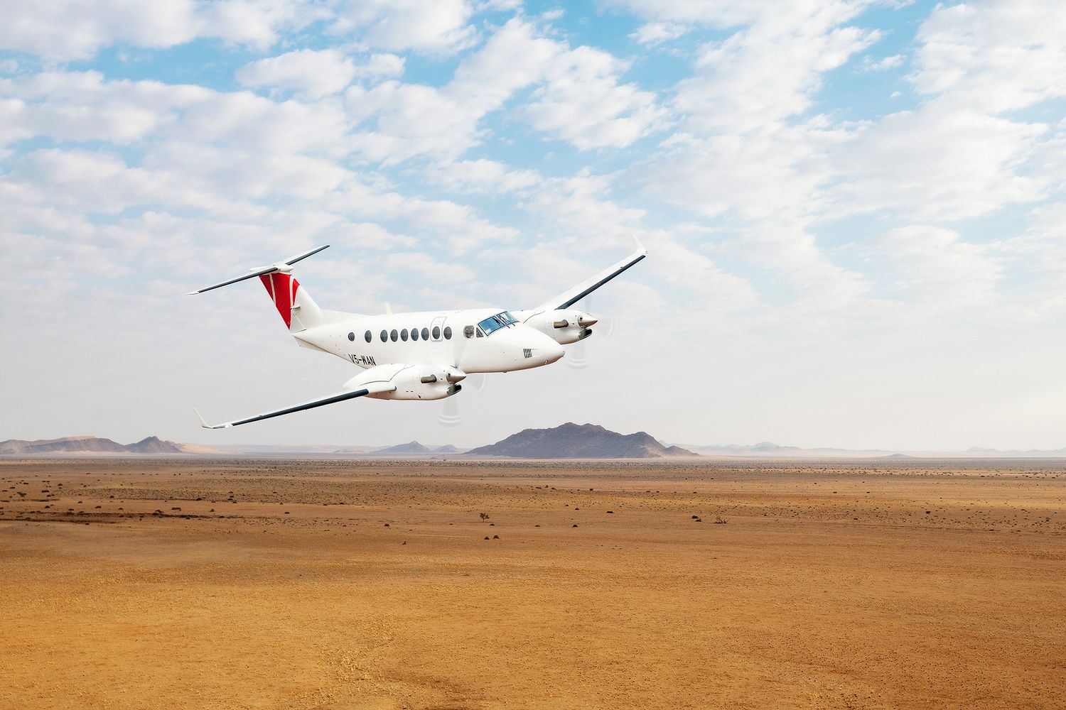A white commercial airplane flies low over a vast arid desert landscape with distant mountains under a partly cloudy sky.