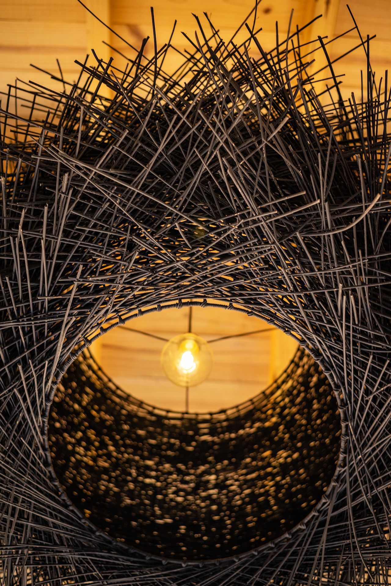 Artistic woven rattan lamp with exposed Edison bulb glowing warmly inside a dark intricately twisted wire nest sculpture.