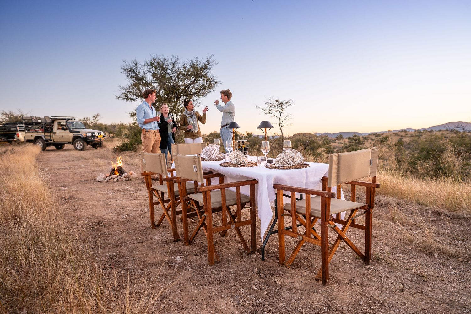 Outdoor bush dinner setup with director chairs and white tablecloth in African savanna at sunset with guests celebrating.