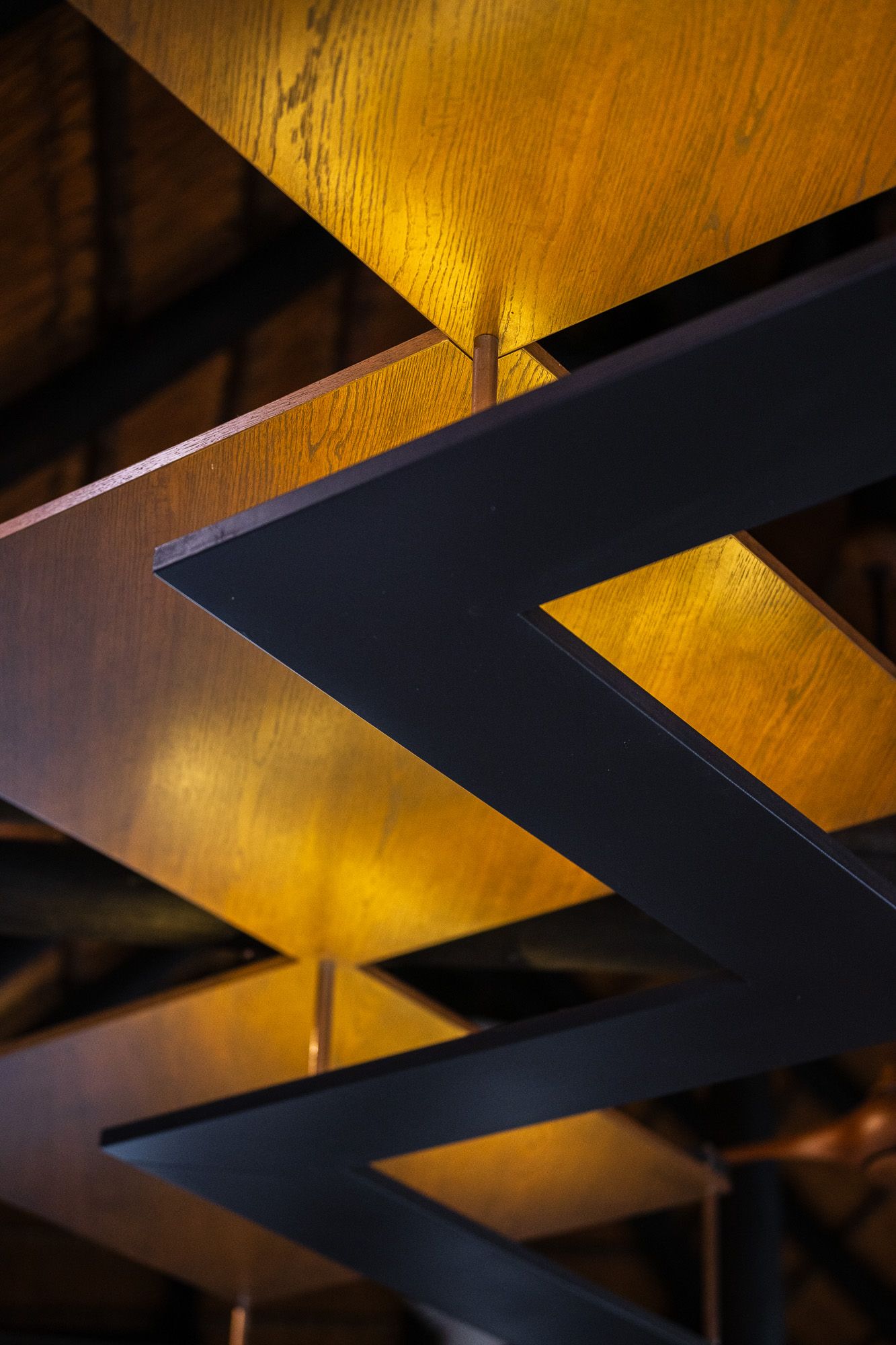 Close-up of a modern ceiling with dark metal grid framework and warm golden wood panels illuminated by hidden lights.