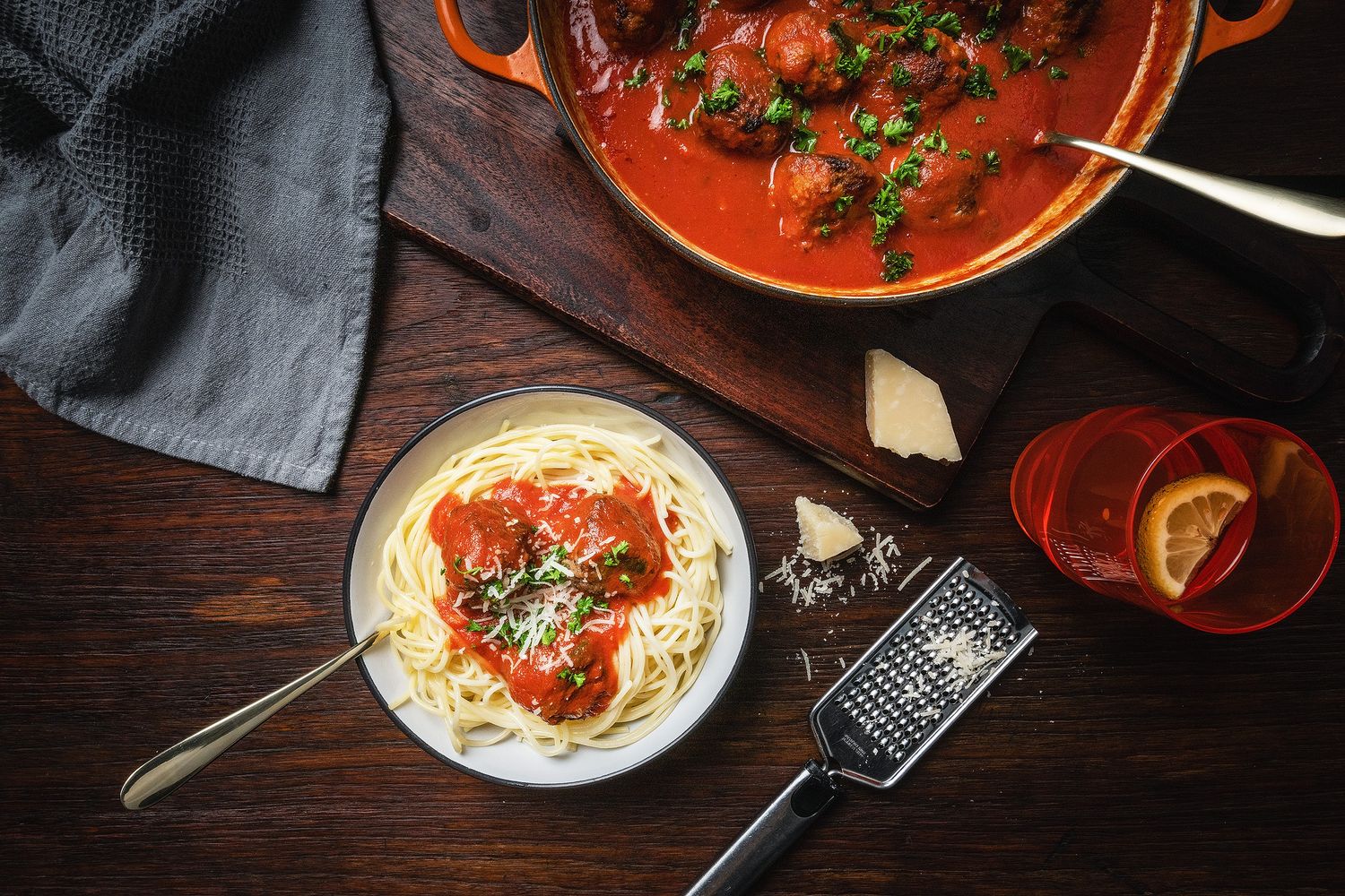 Spaghetti with tomato sauce and meatballs in a white bowl beside a large red cooking pan on a dark wood table.
