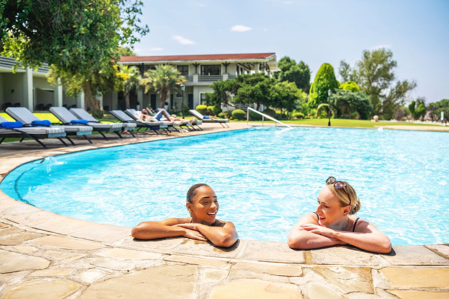 Two women relaxing at a resort swimming pool, leaning on the edge with lounge chairs and tropical greenery in the background.