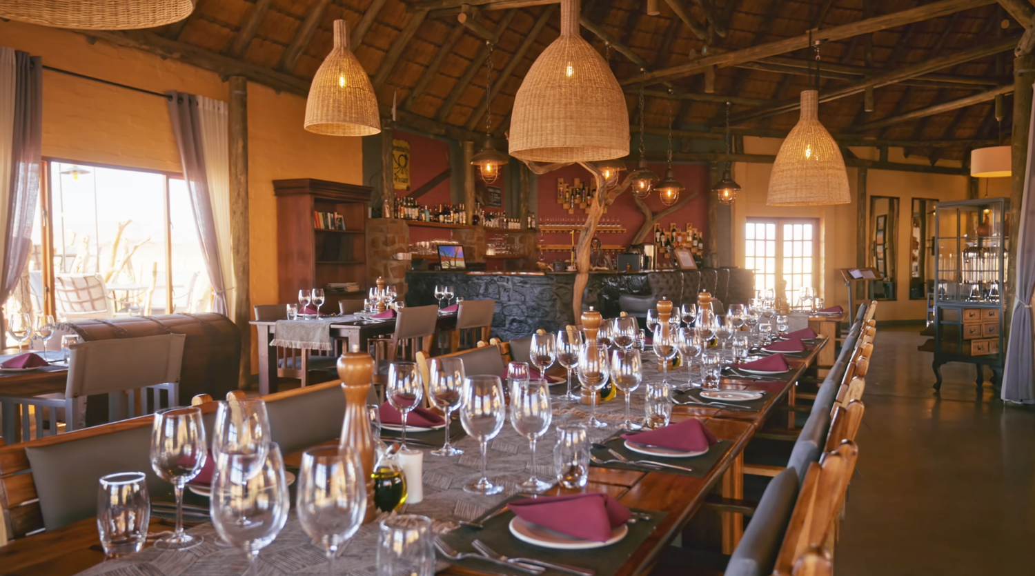 Elegant safari lodge dining room with long wooden tables set with wine glasses and pink napkins under woven pendant lights.
