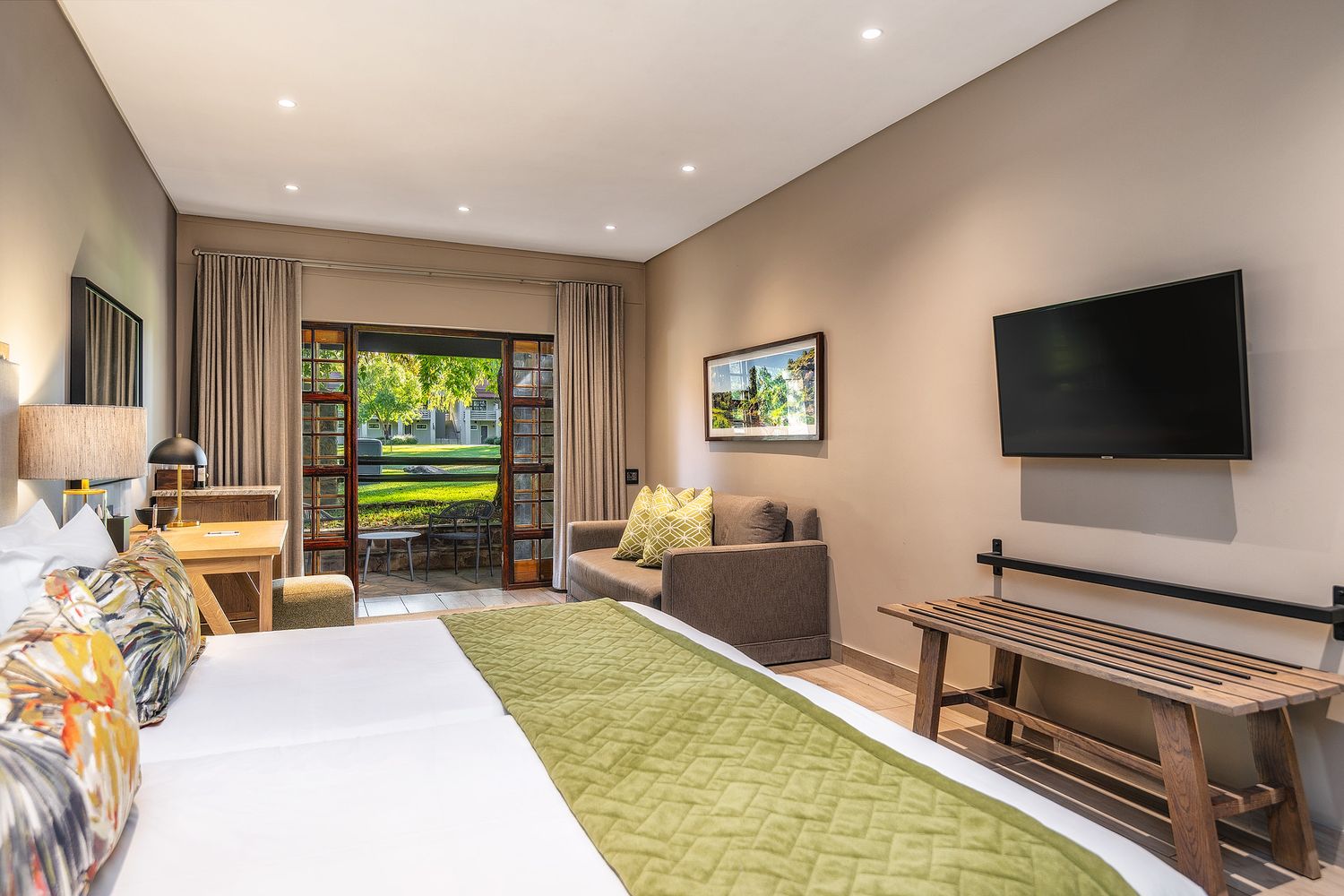 Bright hotel room with king bed, green runner, wall-mounted TV, seating area, and sliding doors opening to a lush garden view.