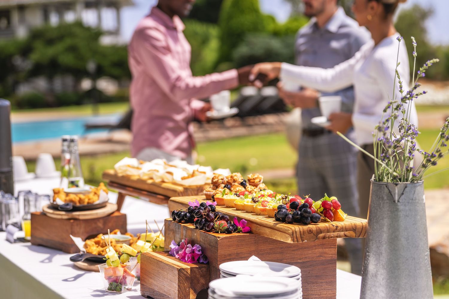 Elegant outdoor buffet table with assorted pastries, fruits, and charcuterie, as guests mingle at a sunny catered event.