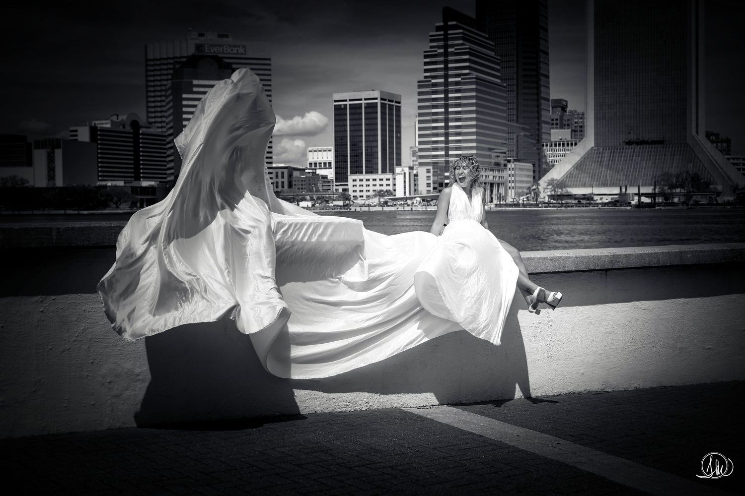 Black and white photo of a woman in flowing white gown sitting outdoors with city skyline in background.
