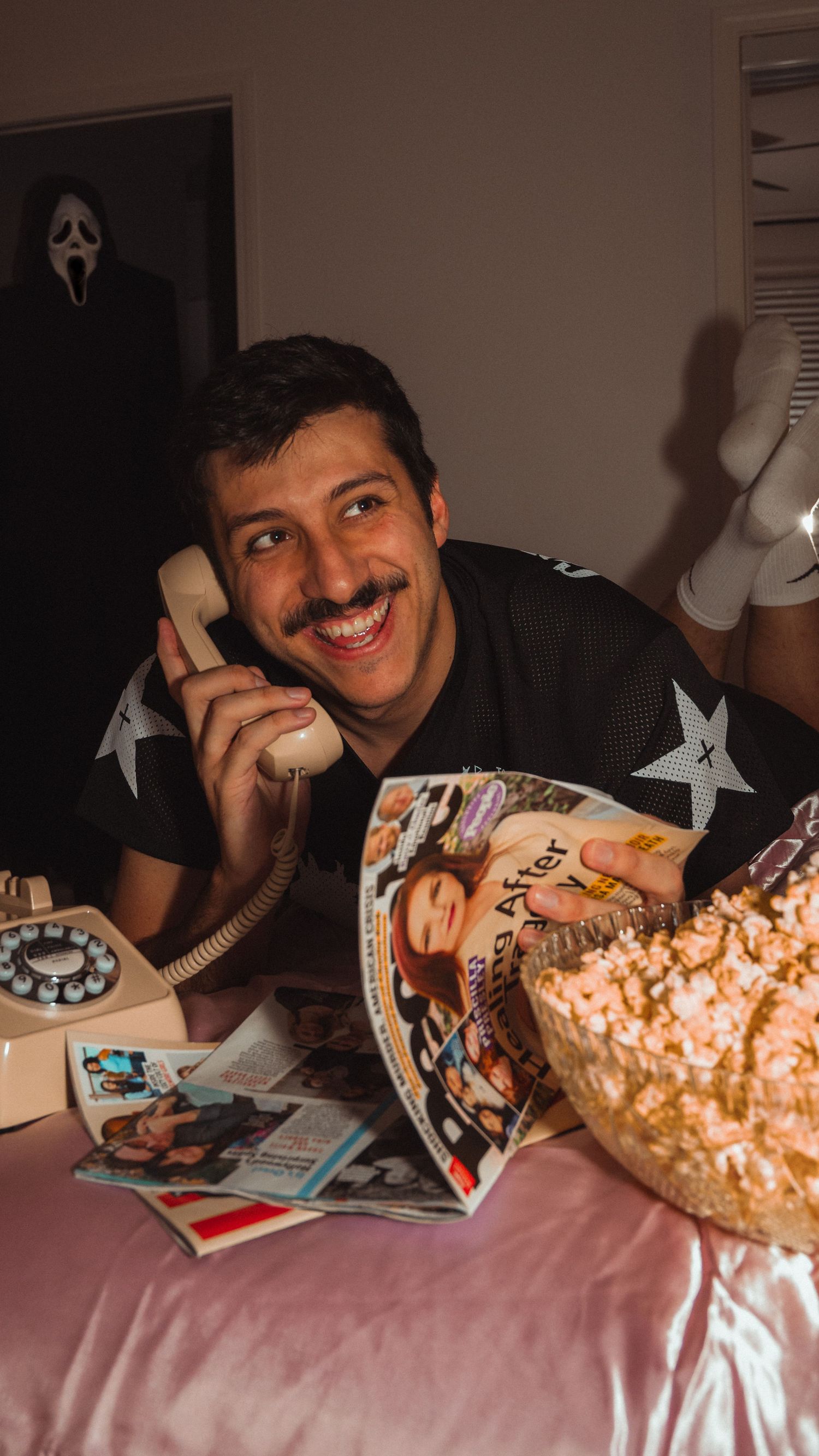 Smiling man with mustache holding retro phone, surrounded by magazines and popcorn, with Ghostface mask in background.