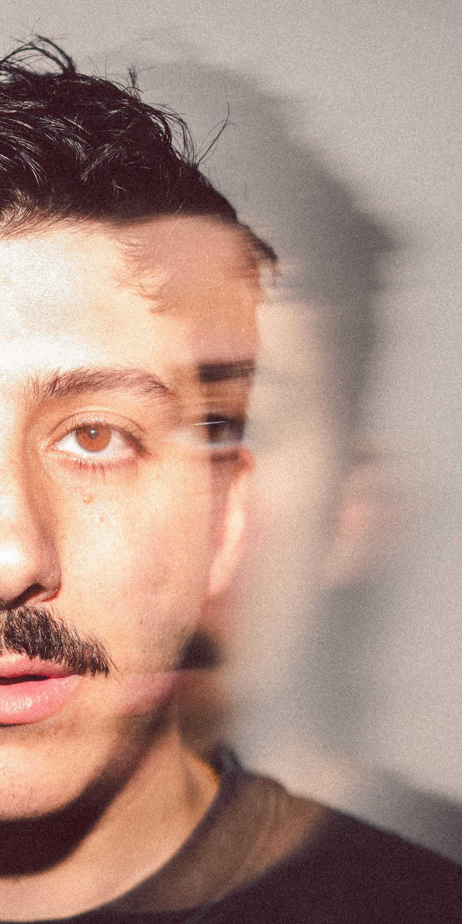 Close-up double exposure portrait of a young man with a mustache and dark hair against a gray background.