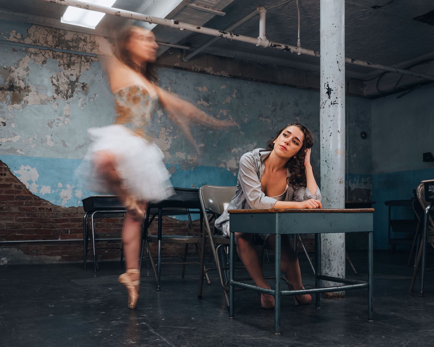 A ballerina in a white tutu dances gracefully while a woman sits at a desk in a rustic, weathered room.