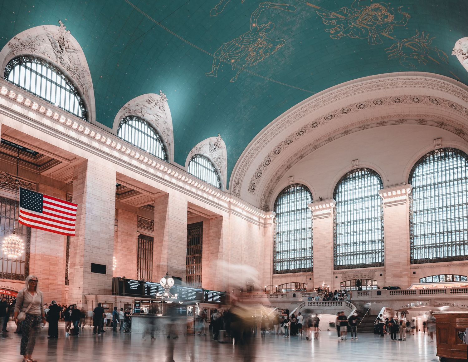 Grand Central Terminal main concourse with American flag, arched windows, and blurred commuters under teal ceiling.